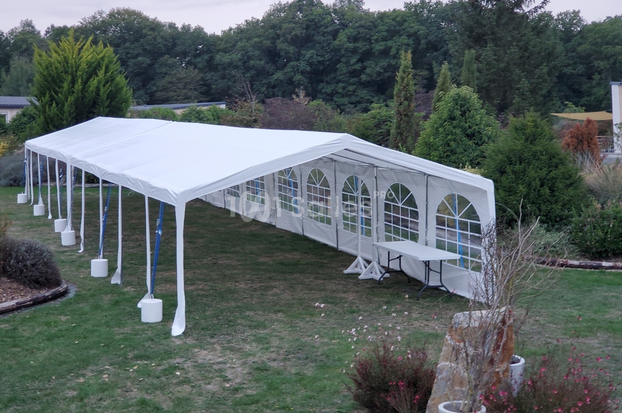 Tente blanche rectangulaire montée dans un jardin, avec des fenêtres en plastique transparent sur les côtés.