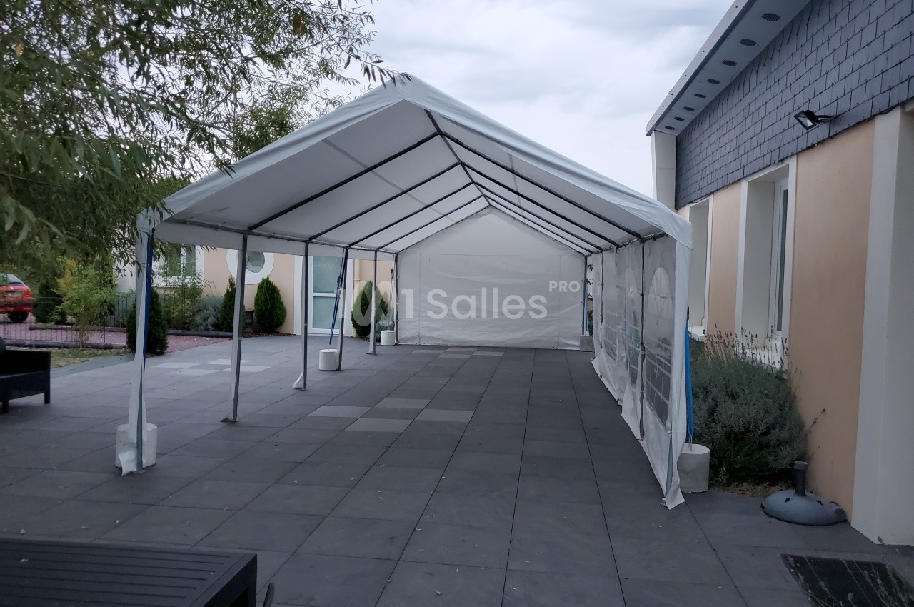 Tente blanche installée sur une terrasse pavée, adossée à un bâtiment beige sous un ciel nuageux.