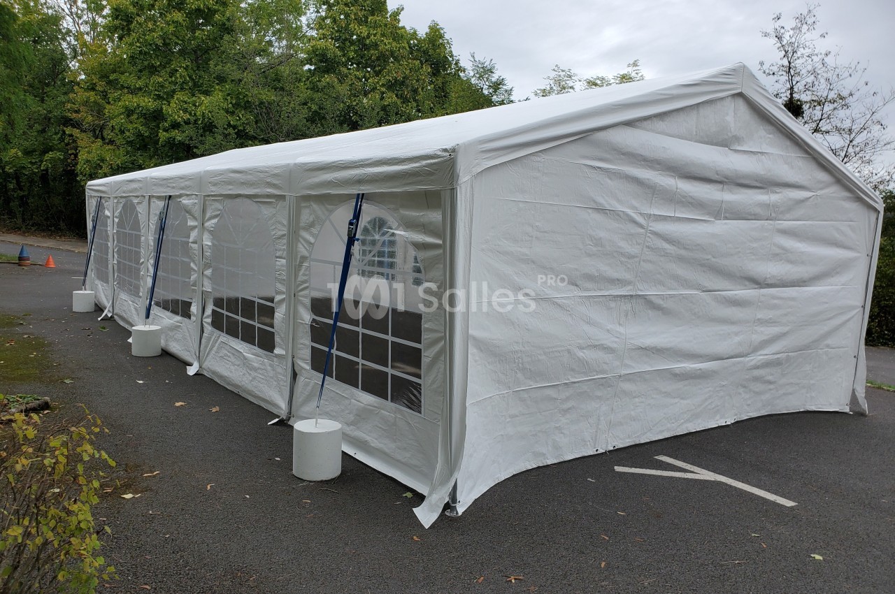 Grande tente blanche rectangulaire installée sur un parking, entourée de végétation et de barrières de sécurité.