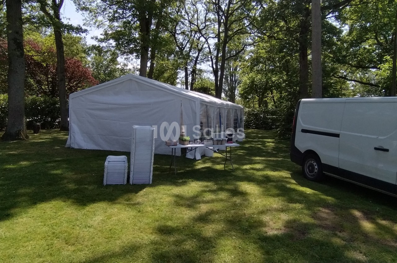 Tente blanche installée dans un jardin verdoyant, avec des tables, des chaises empilées et une camionnette stationnée à côté.