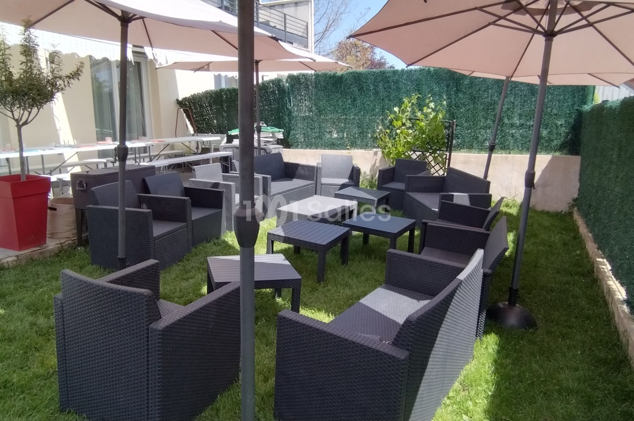 Salon de jardin en résine noire avec tables et parasols, installé sur une pelouse entourée de haies vertes.