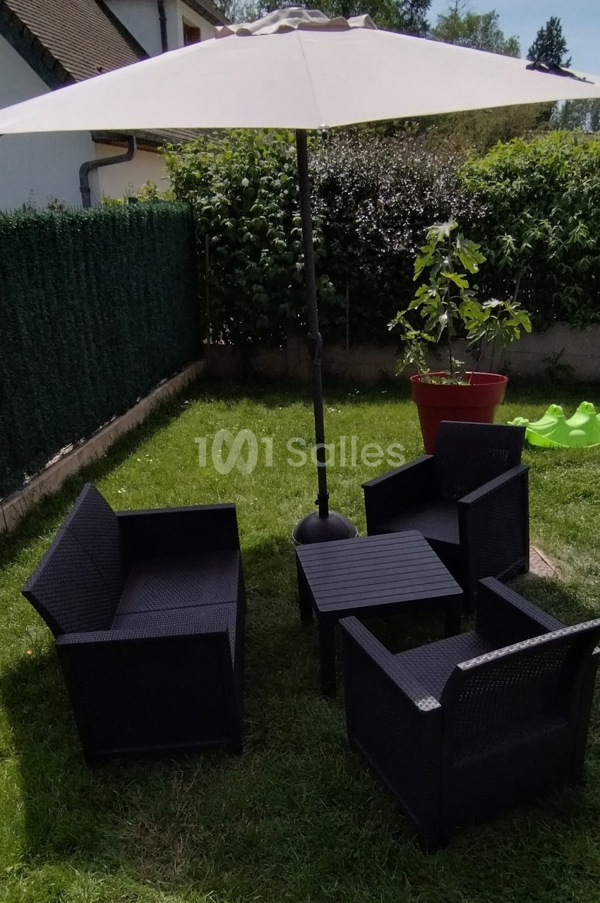 Salon de jardin en résine noire avec parasol blanc, installé sur une pelouse près d'une haie et d'un pot de fleurs.