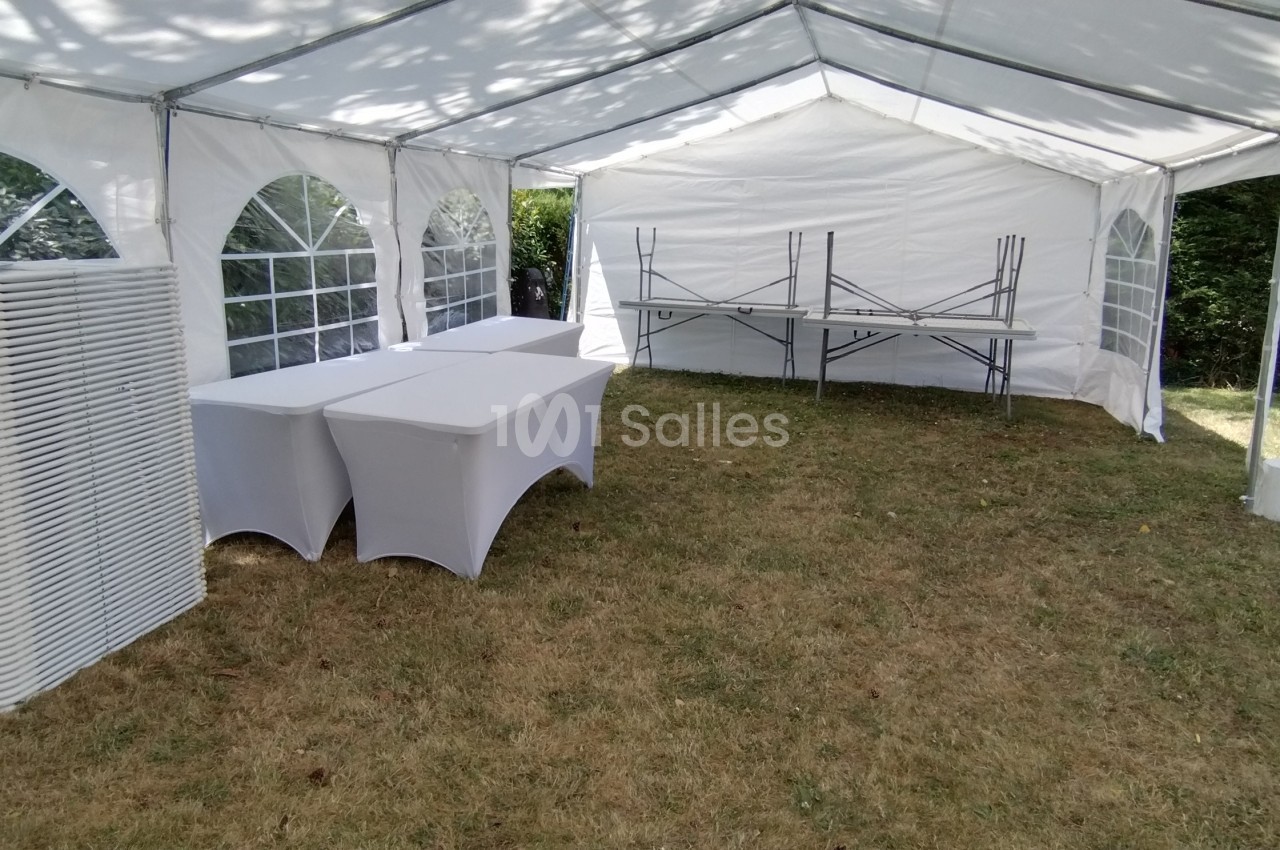 Tente blanche montée dans un jardin, avec des tables pliantes et des chaises empilées sur un sol en herbe.