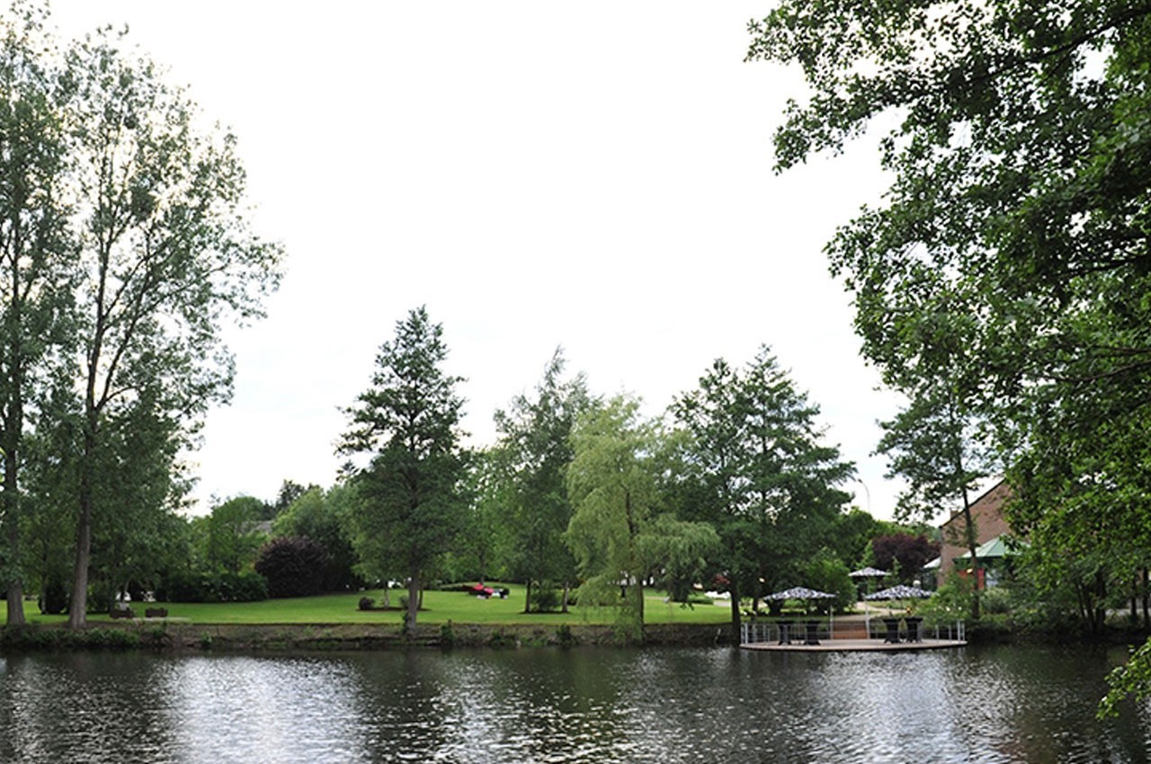 Vue d'un parc verdoyant avec des arbres, une pelouse et une rivière au premier plan, sous un ciel clair.