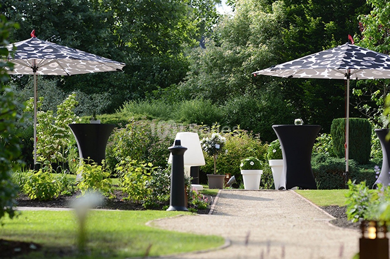 Allée de jardin bordée de verdure, avec tables hautes noires et parasols à motifs noirs et blancs.