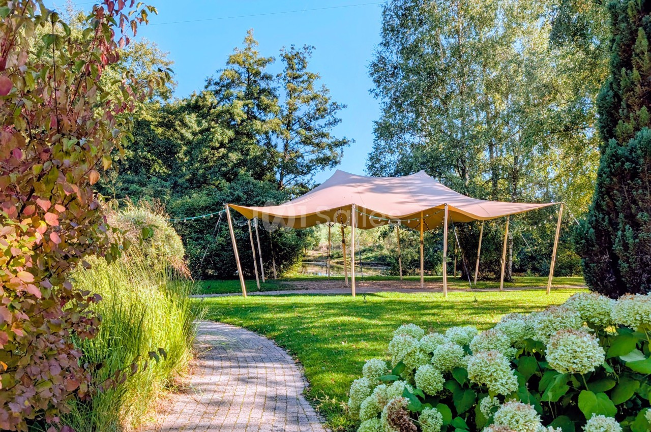 Tente beige montée dans un jardin verdoyant, entourée d'arbustes, de fleurs et d'arbres sous un ciel bleu.