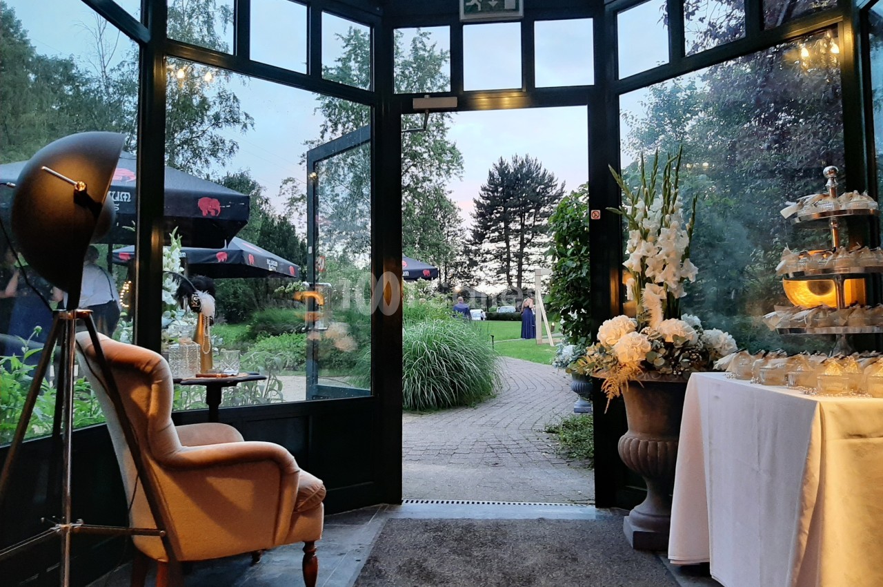 Vue depuis une véranda vitrée donnant sur un jardin arboré, avec fauteuil, table décorée et éclairage tamisé.