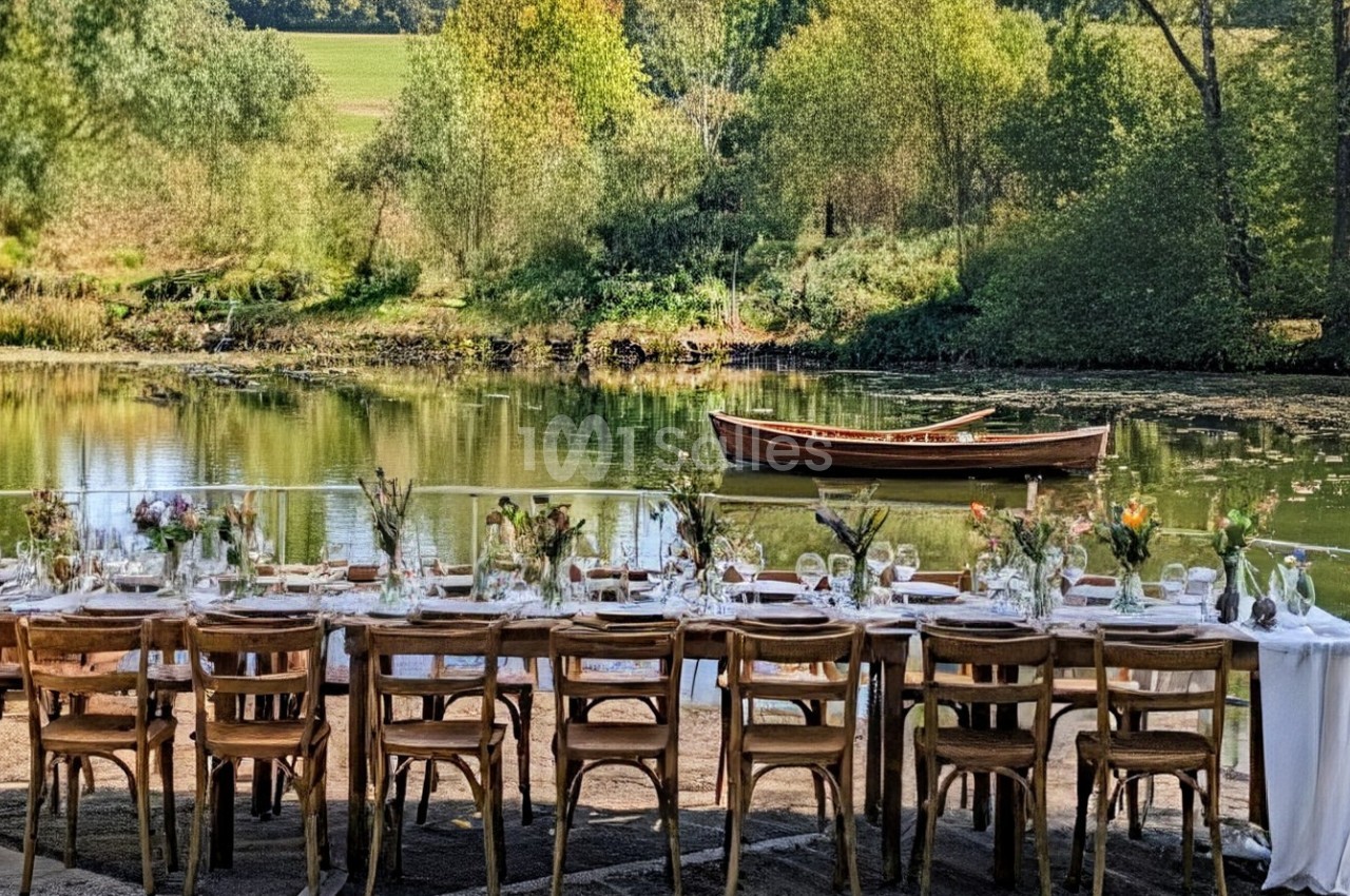 Table dressée en extérieur avec nappes blanches, chaises en bois, fleurs et vue sur un étang avec une barque.