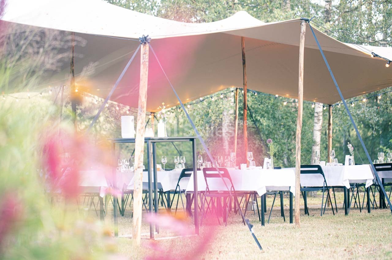 Tables dressées sous une tente beige dans un jardin, avec des fleurs floues au premier plan.