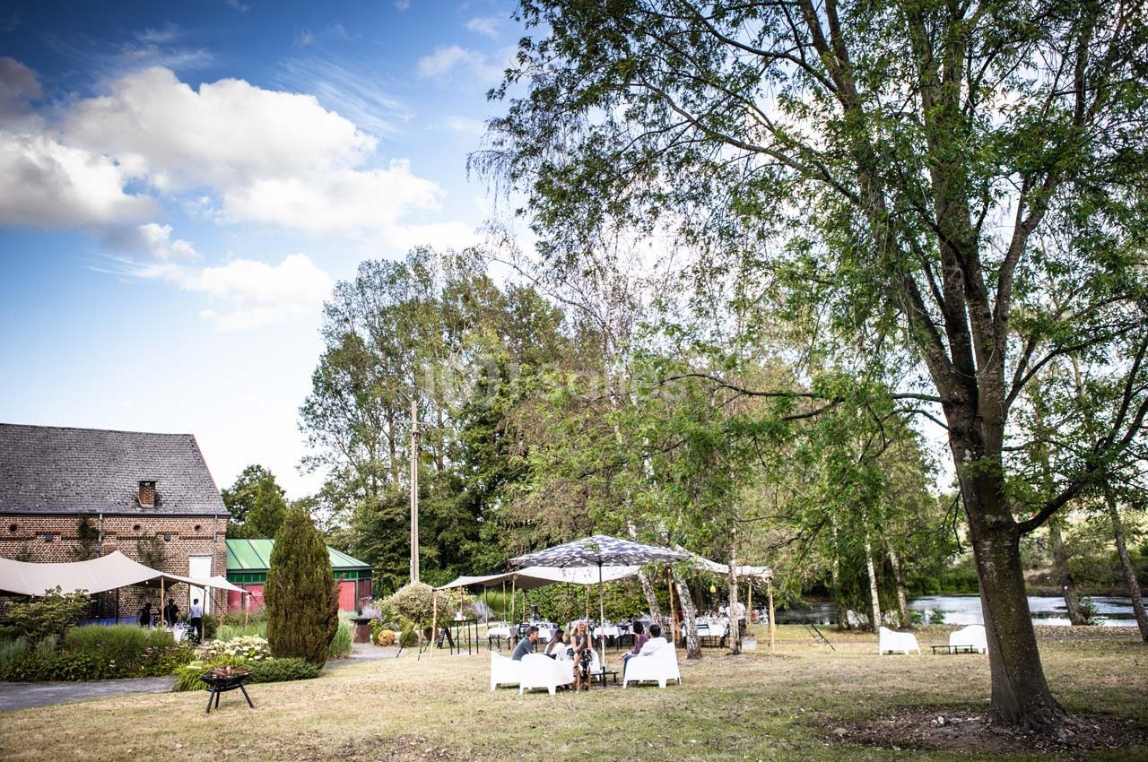 Jardin avec des tables et chaises sous des parasols, près d'une rivière et d'un bâtiment en pierre, entouré d'arbres.