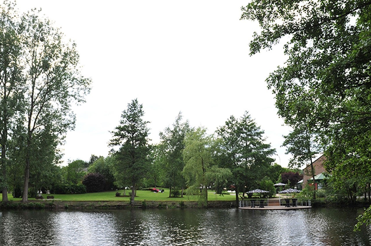 Vue d'un parc verdoyant avec des arbres, une pelouse et une rivière au premier plan, sous un ciel clair.