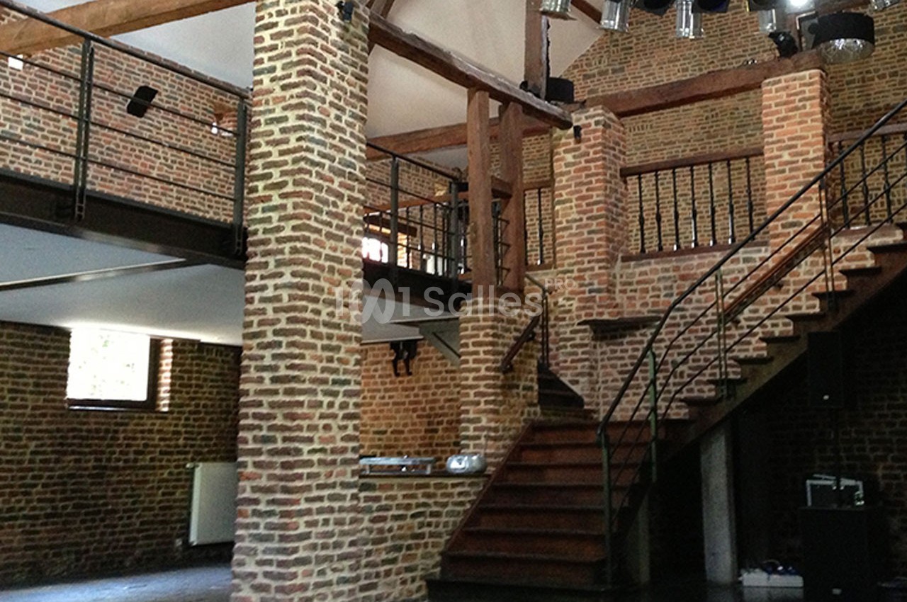 Intérieur d'un bâtiment en briques avec mezzanine, escalier en bois et poutres apparentes.