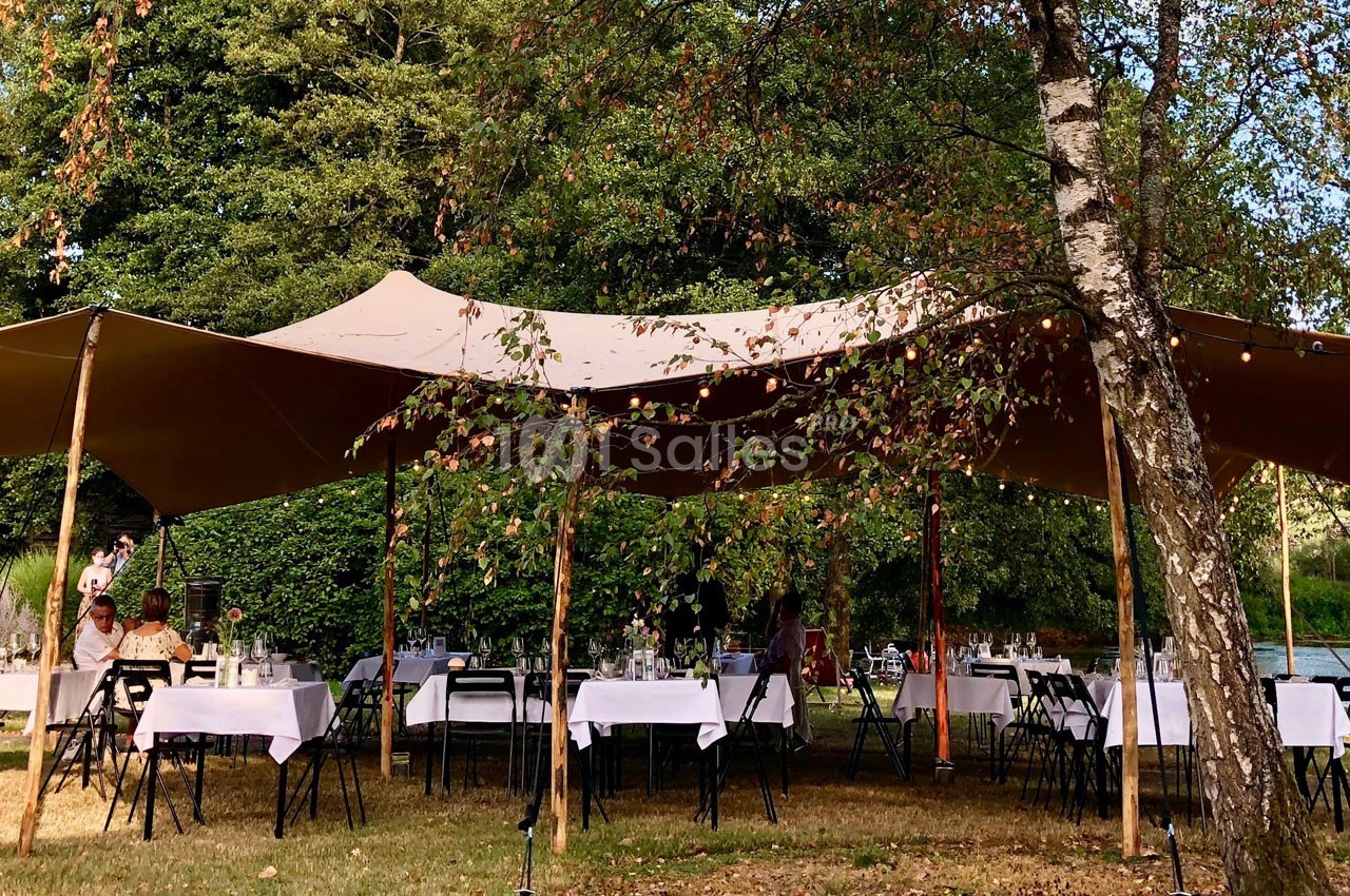 Tables dressées sous une grande tente beige dans un jardin arboré, avec des guirlandes lumineuses suspendues.