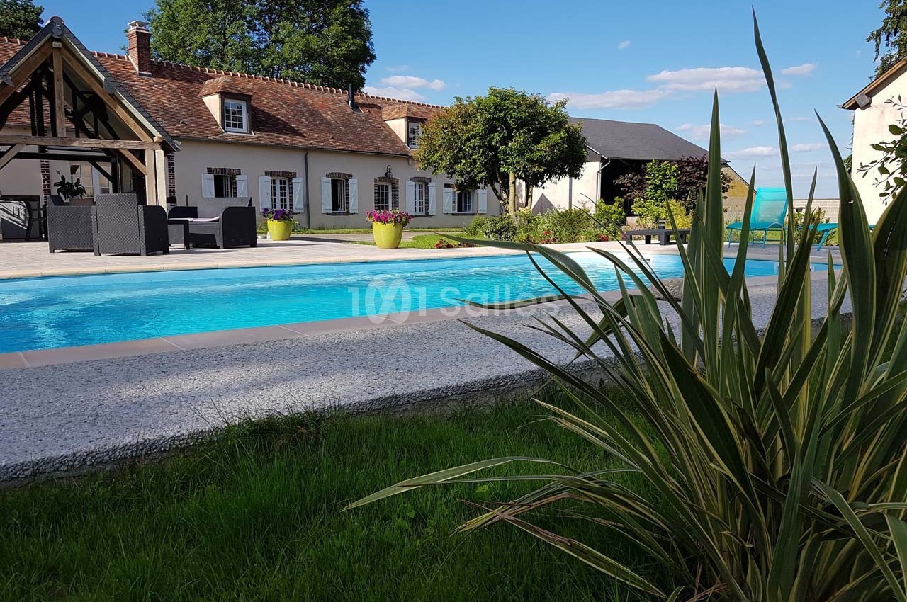 Maison traditionnelle avec piscine extérieure, terrasse aménagée et jardin arboré sous un ciel dégagé.