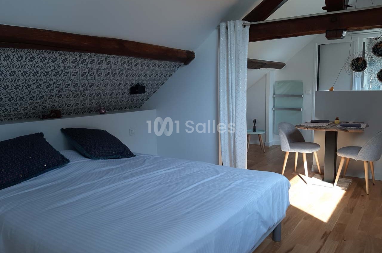 Chambre mansardée avec lit double, table à manger, chaises et poutres apparentes, éclairée par une fenêtre.