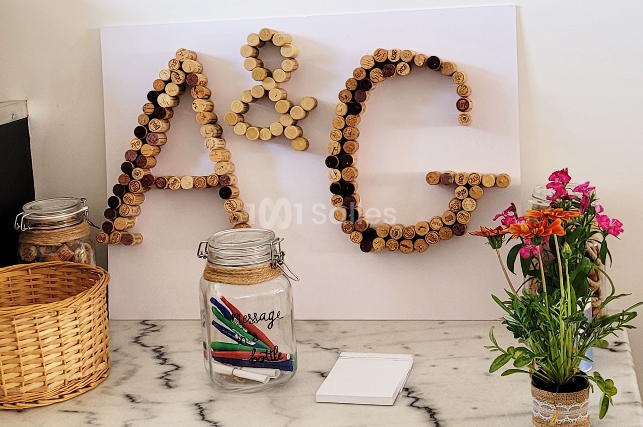 Lettres ’A & G’ formées avec des bouchons en liège, entourées de pots en verre, d'un panier et d'un vase de fleurs.