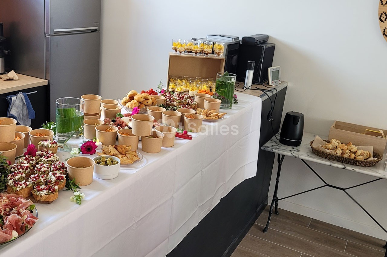 Buffet avec divers plats, verrines, pains et garnitures disposés sur une table blanche dans une salle lumineuse.