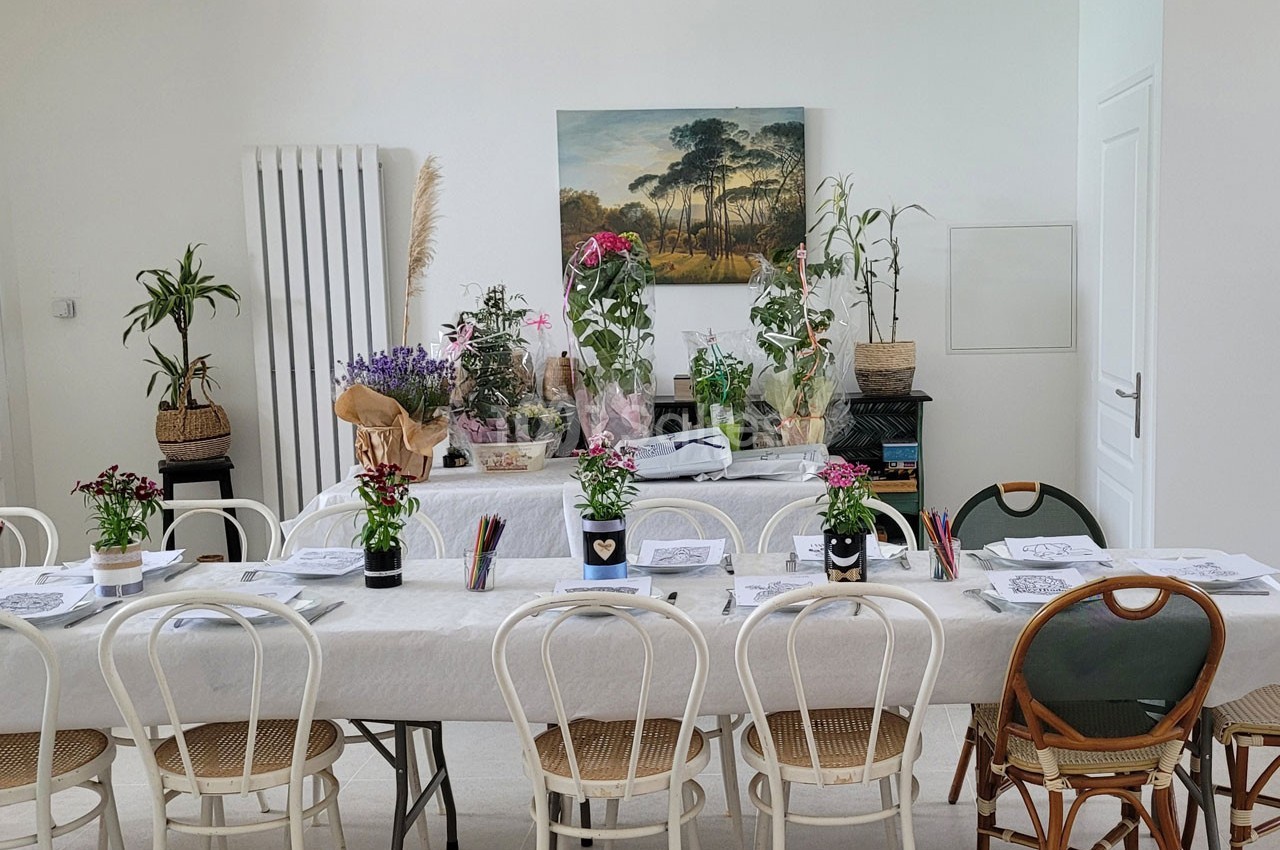 Salle lumineuse avec deux tables décorées de plantes, fleurs et fournitures de dessin, entourées de chaises variées.