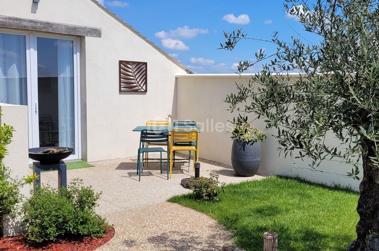 Terrasse extérieure avec table et chaises colorées, barbecue, olivier en pot et pelouse sous un ciel bleu.