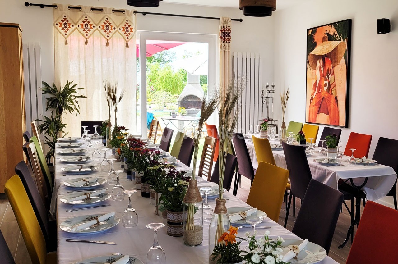Salle lumineuse avec deux longues tables dressées, décorées de fleurs et entourées de chaises colorées.