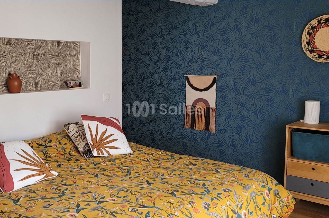 Chambre avec un lit à motif floral, mur bleu texturé, étagère intégrée et décoration murale en tissu et osier.