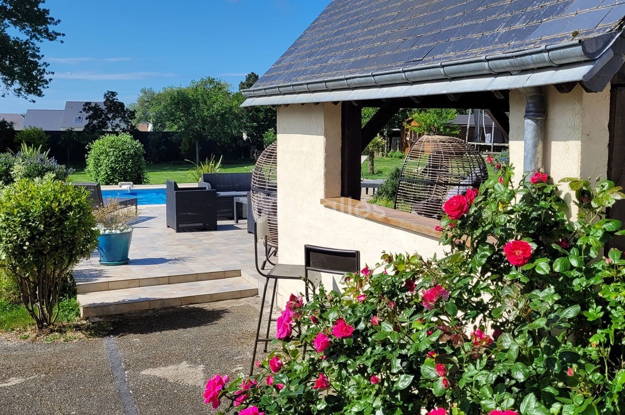 Jardin avec terrasse, piscine, rosiers en fleurs et mobilier d'extérieur sous un ciel dégagé.