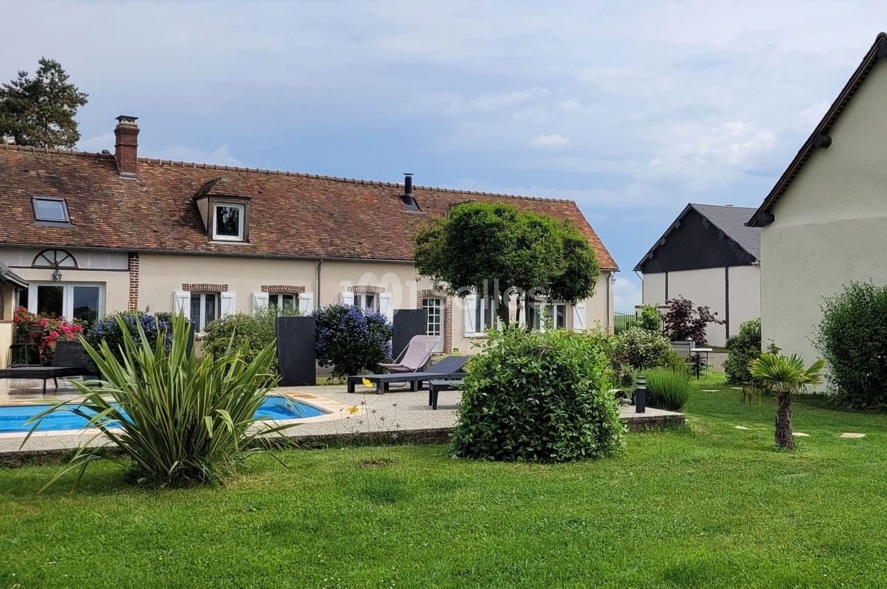 Maison traditionnelle avec piscine, terrasse aménagée et jardin verdoyant sous un ciel partiellement nuageux.