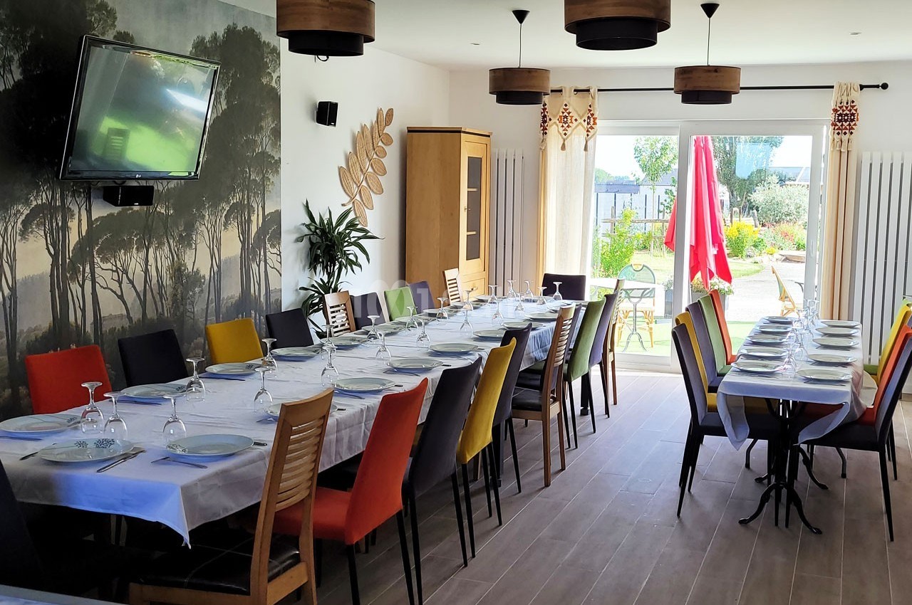 Salle à manger lumineuse avec de longues tables dressées, des chaises colorées et une vue sur un jardin extérieur.