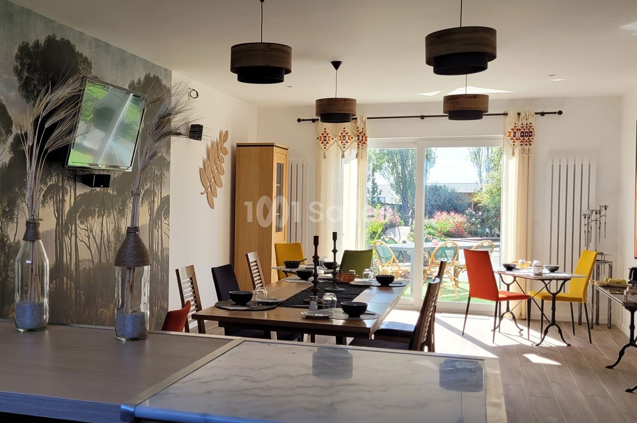 Salle à manger lumineuse avec table dressée, chaises colorées, baies vitrées donnant sur un jardin ensoleillé.