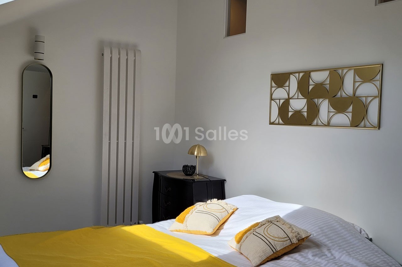 Chambre lumineuse avec lit double, couvre-lit jaune, coussins assortis, miroir, radiateur et décoration murale dorée.