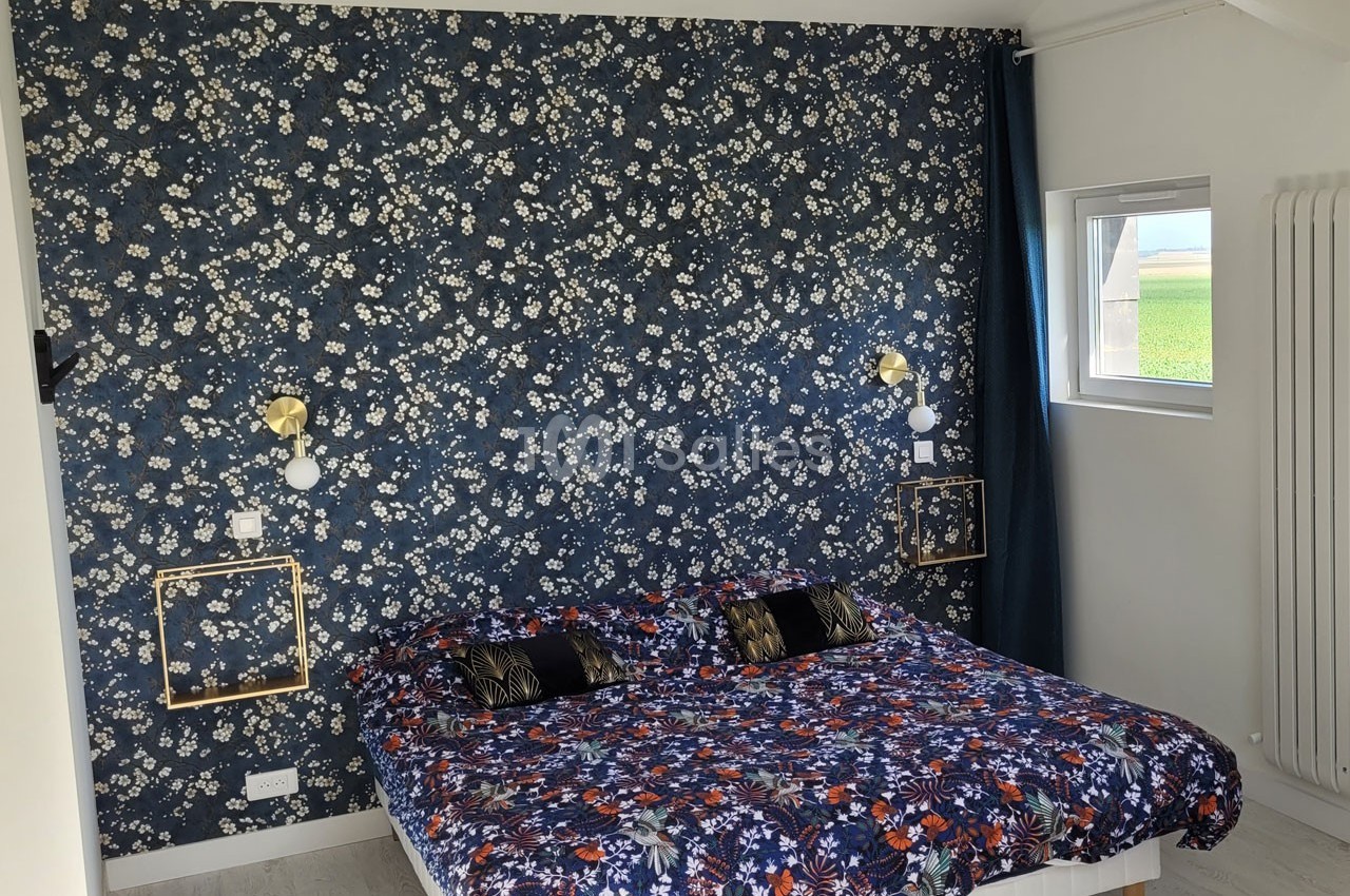 Chambre avec un lit double, mur tapissé de motifs floraux bleus, luminaires dorés et fenêtre laissant entrer la lumière.