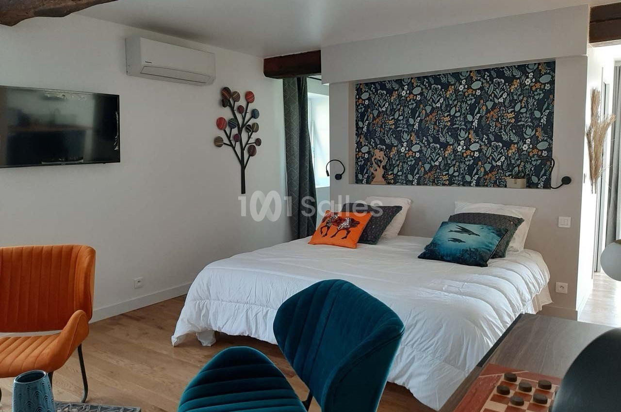 Chambre lumineuse avec lit double, décor floral sur le mur, fauteuils colorés et télévision fixée au mur.