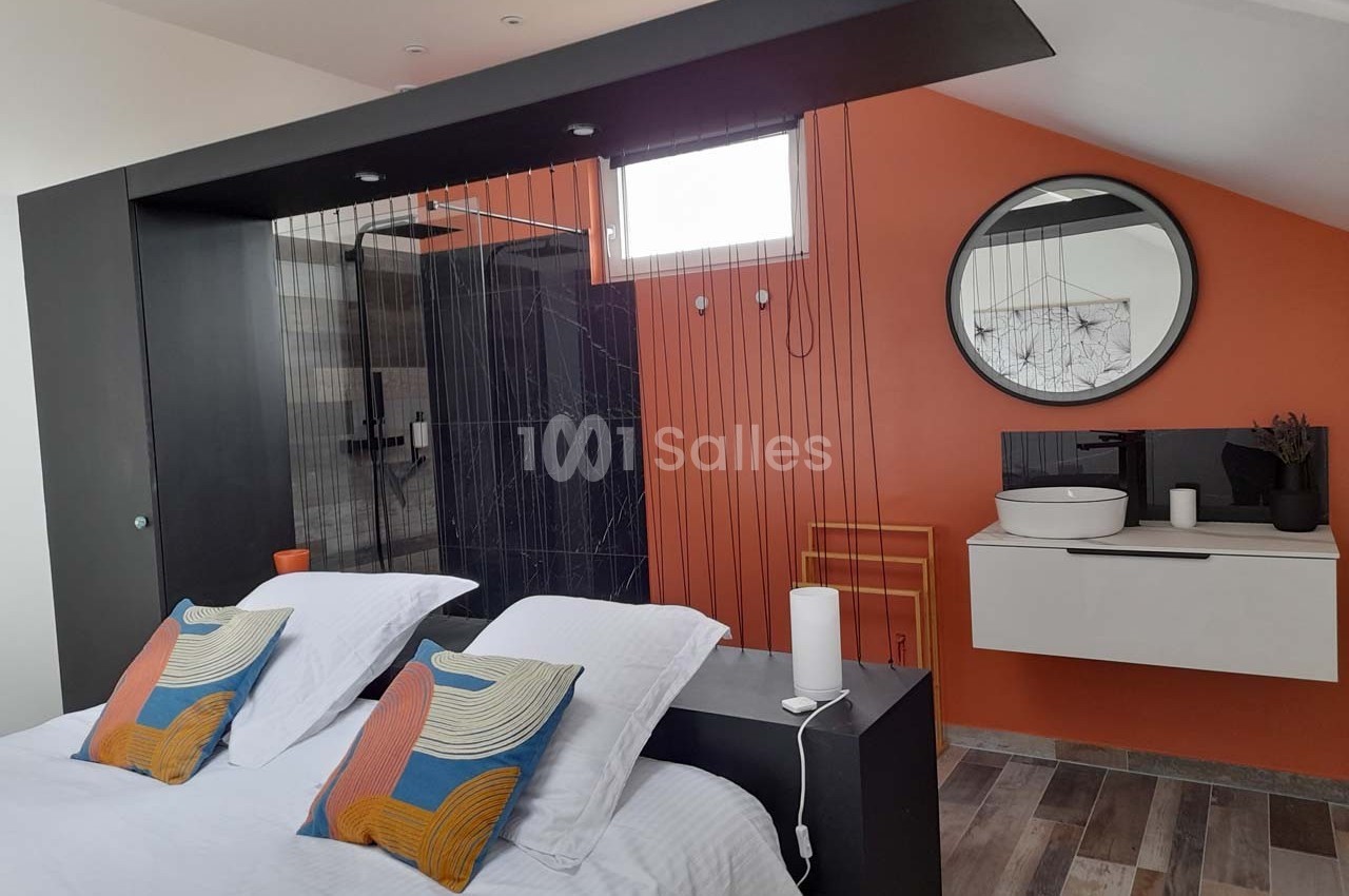 Chambre moderne avec lit double, cloison ajourée, mur orange, salle de bain attenante et lavabo sur meuble suspendu.