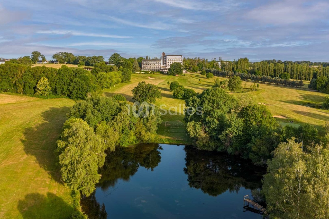 Vue aérienne d'un paysage verdoyant avec un étang au premier plan et un grand bâtiment au loin entouré d'arbres.
