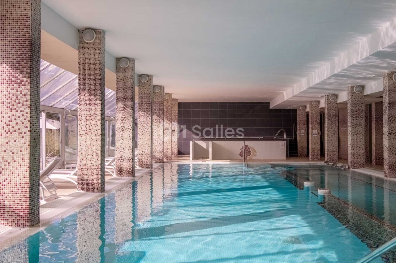 Piscine intérieure lumineuse entourée de colonnes carrelées, avec chaises longues et grandes baies vitrées.