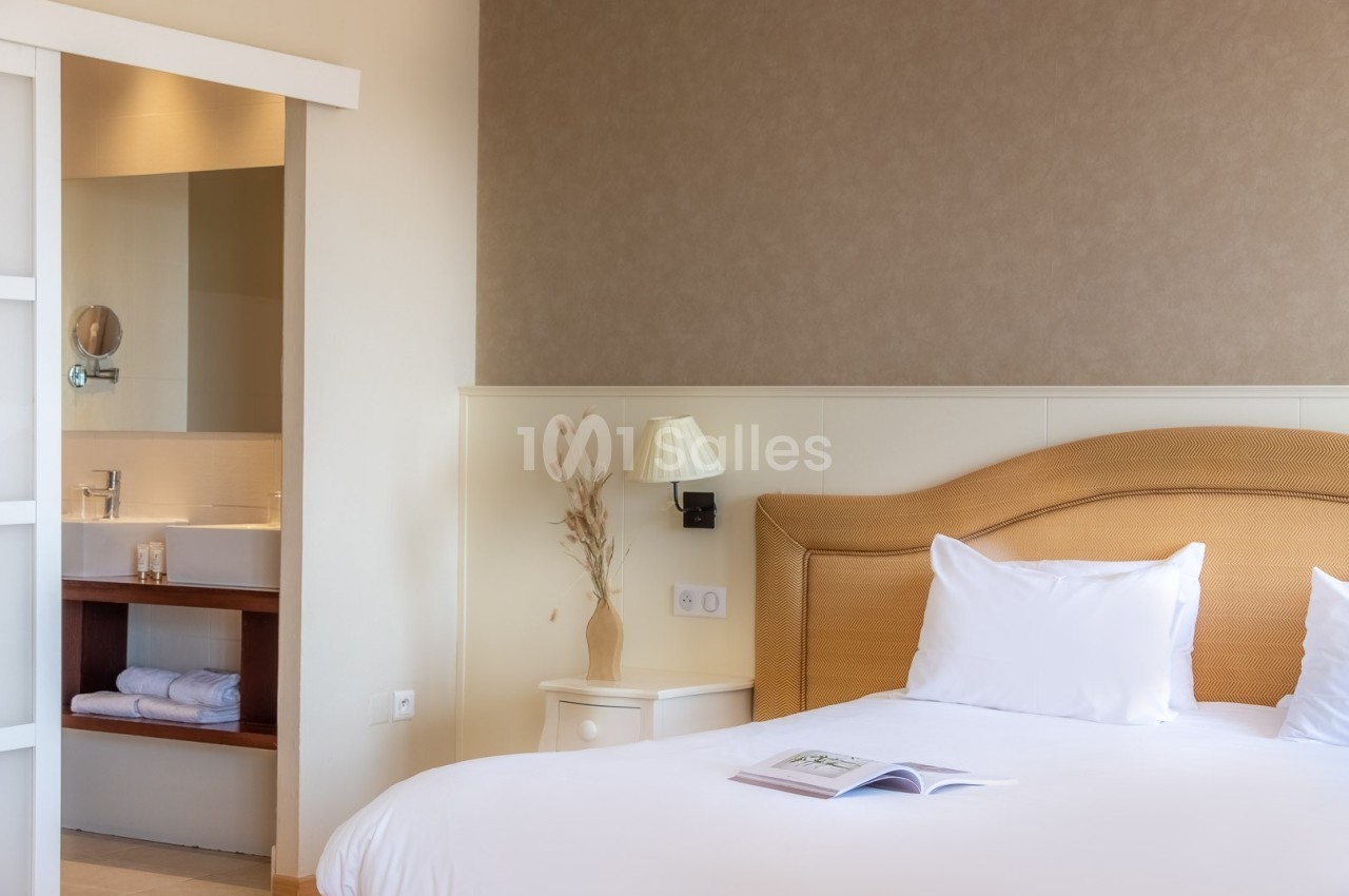 Chambre lumineuse avec lit double, table de chevet, lampe et vue sur une salle de bain avec lavabo moderne.