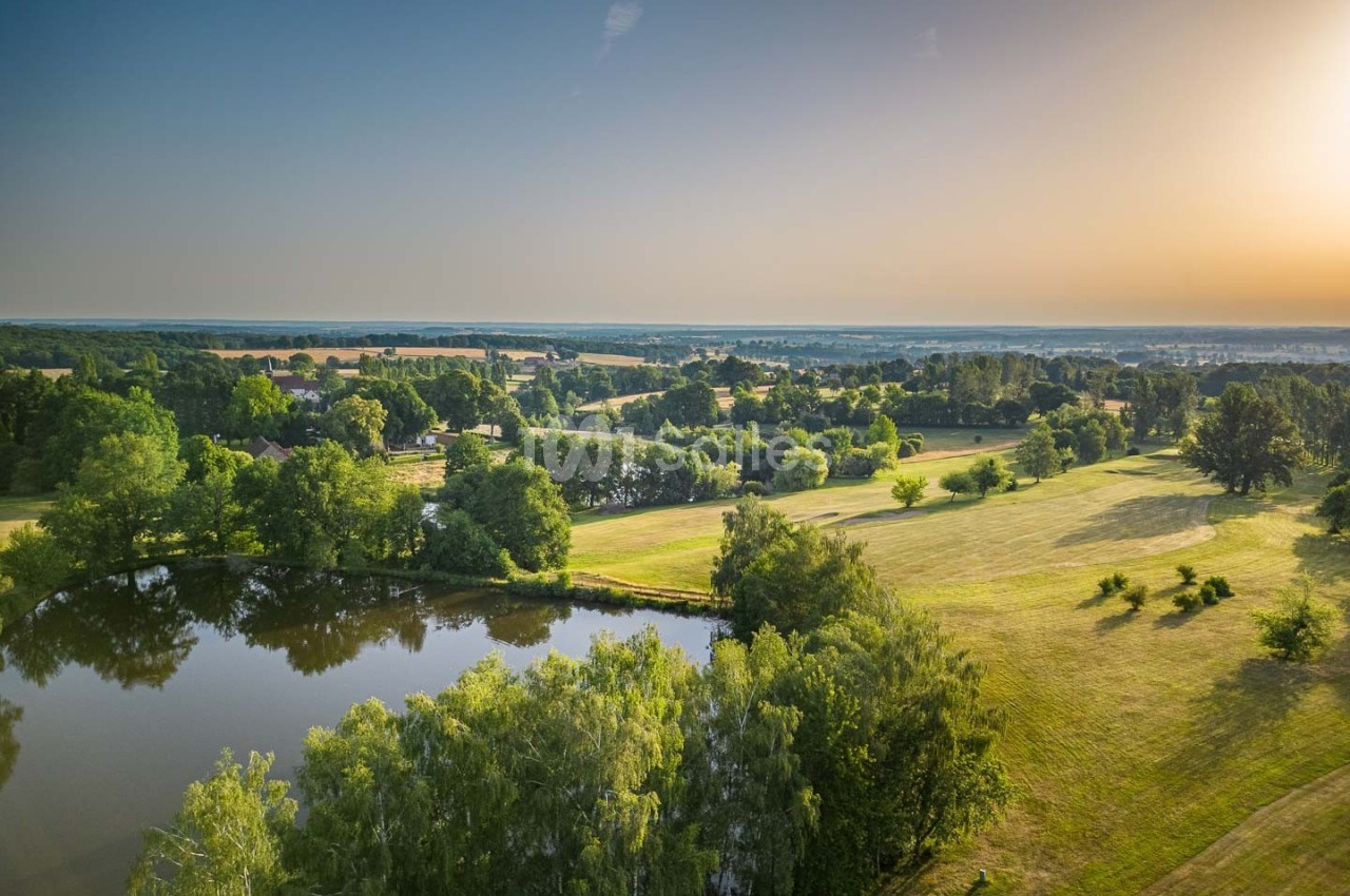 Vue aérienne d'un paysage rural avec un étang entouré d'arbres et des champs s'étendant à l'horizon sous un ciel dégagé.