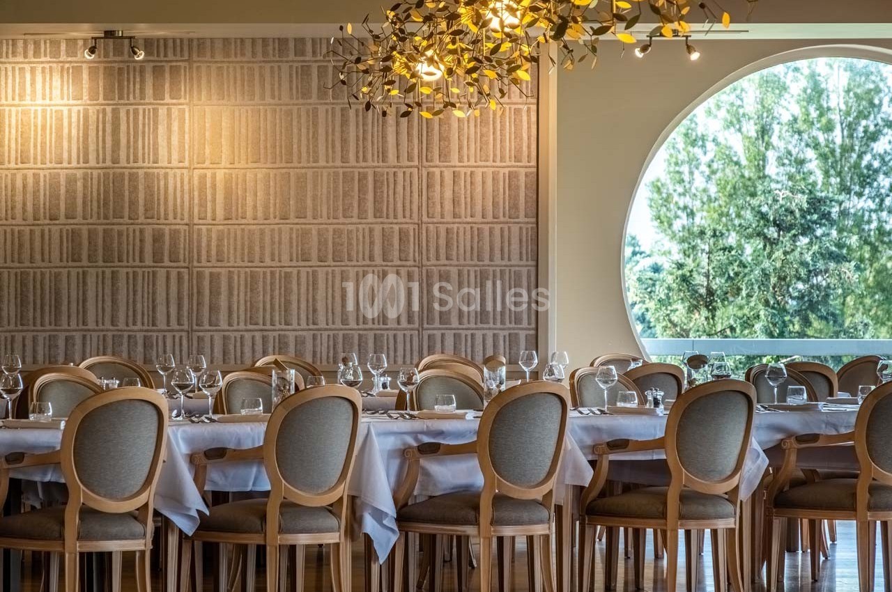 Salle de restaurant élégante avec tables dressées, chaises en bois, mur texturé et grande fenêtre ronde donnant sur des…