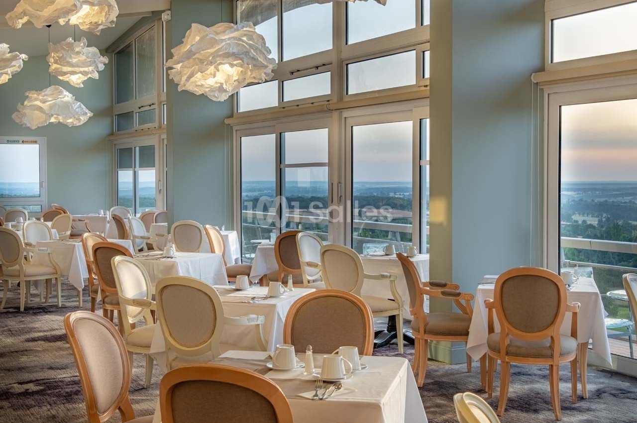 Salle de restaurant lumineuse avec grandes baies vitrées, tables dressées et vue sur un paysage dégagé.