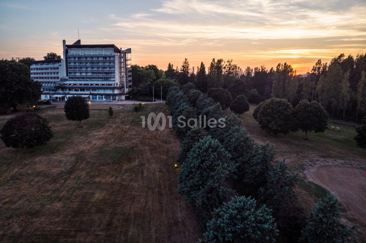 Vue aérienne d'un bâtiment moderne entouré d'arbres et d'une pelouse, avec un coucher de soleil en arrière-plan.