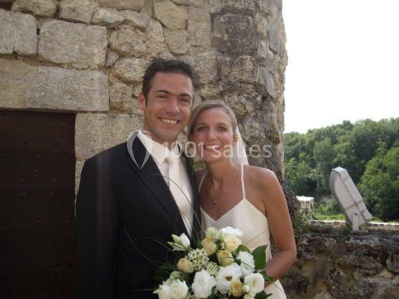 Un couple souriant en tenue de mariage pose devant un mur en pierre, tenant un bouquet de fleurs blanches.