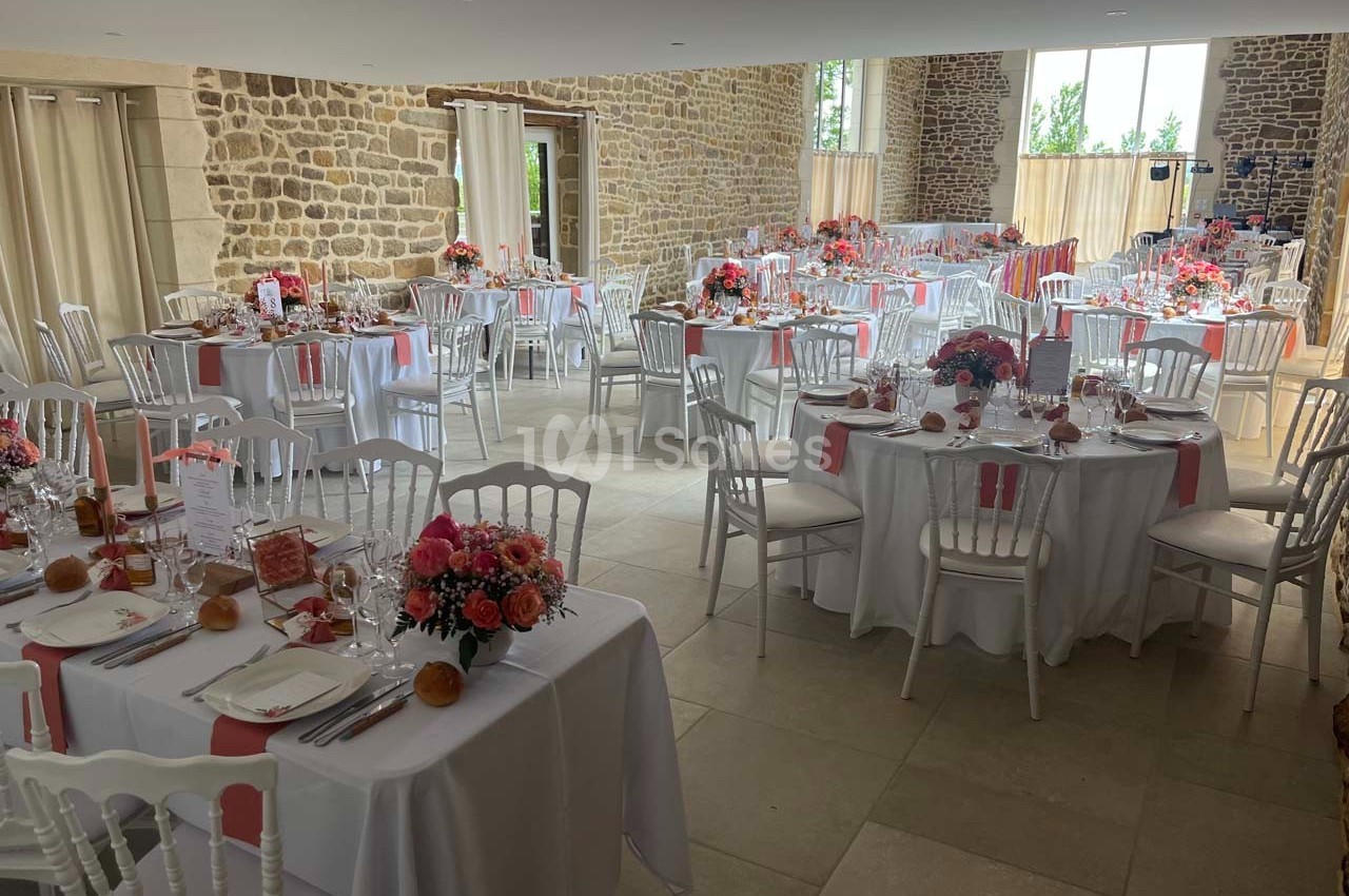 Salle de réception décorée avec des tables rondes, nappes blanches, chaises blanches et arrangements floraux colorés.