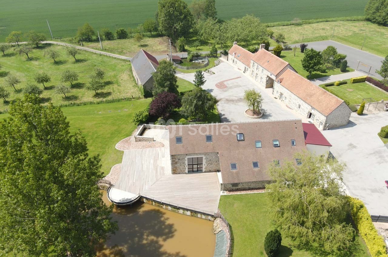 Vue aérienne d'un domaine rural avec bâtiments en pierre, cour pavée, étang et champs verdoyants en arrière-plan.