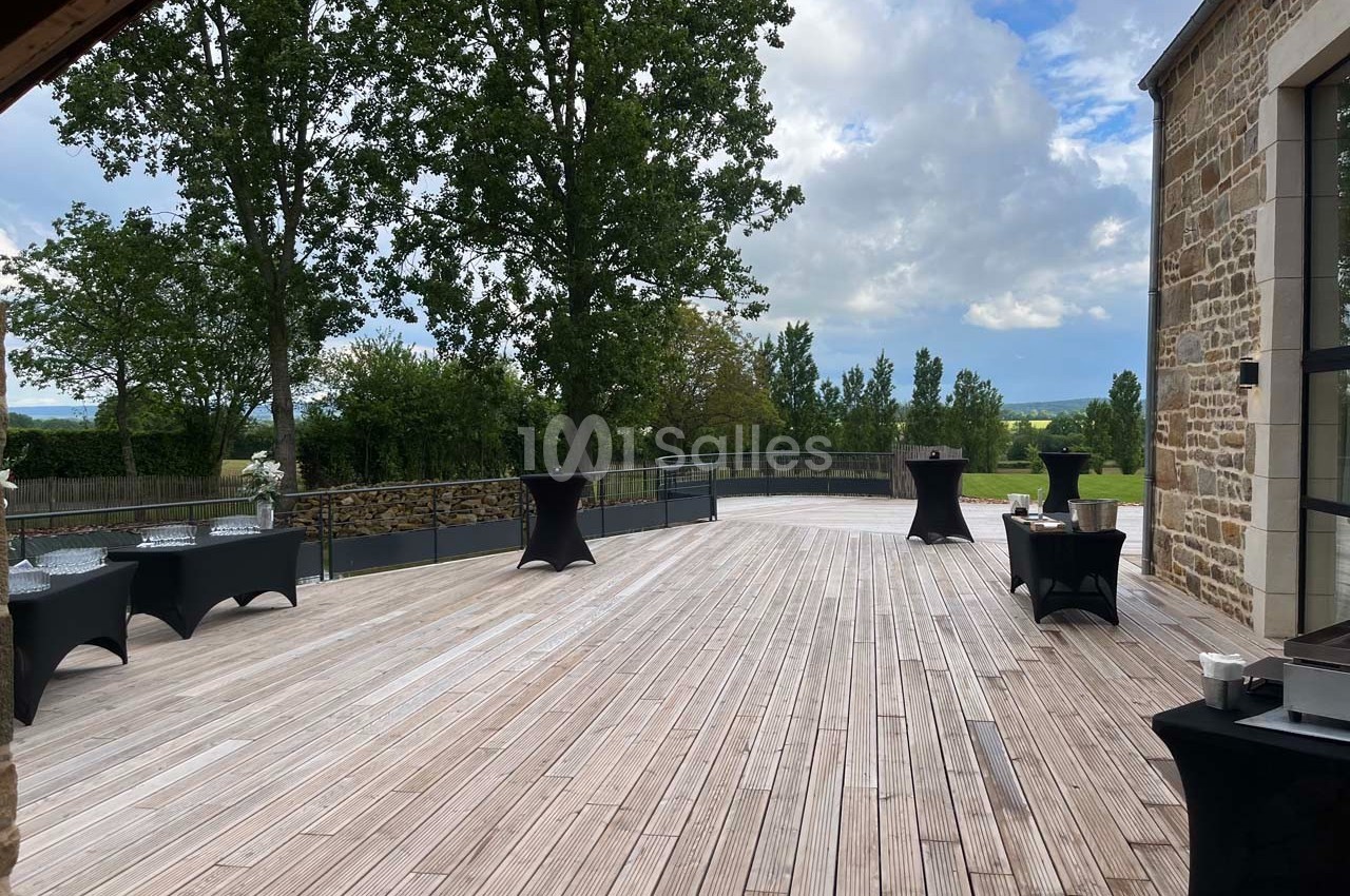 Terrasse en bois avec tables hautes noires, bordée d'arbres et offrant une vue dégagée sur la campagne.