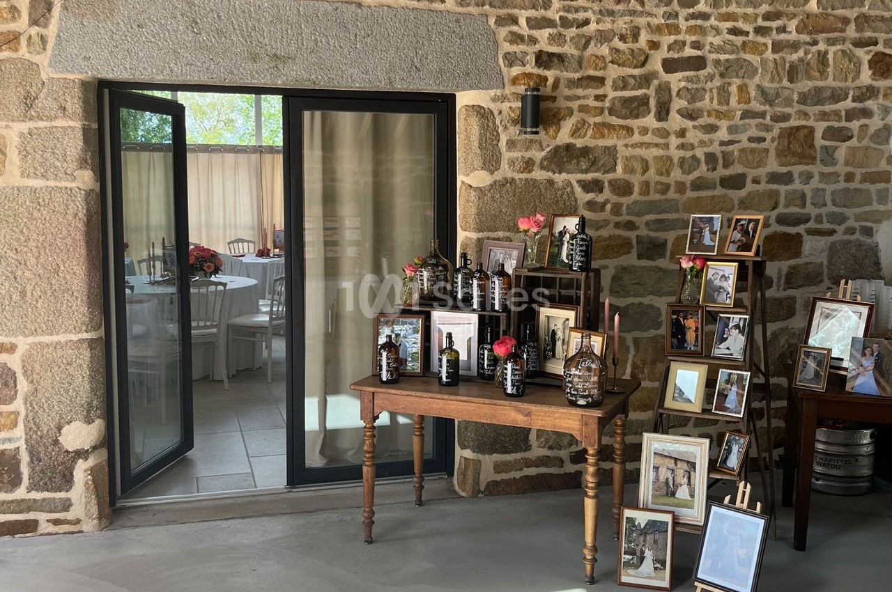 Table en bois avec des bouteilles décoratives et des fleurs, entourée de cadres photo, devant un mur en pierre et une porte…