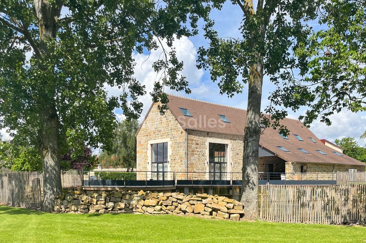 Maison en pierre avec toit en pente, entourée d'arbres et d'une pelouse, sous un ciel partiellement nuageux.