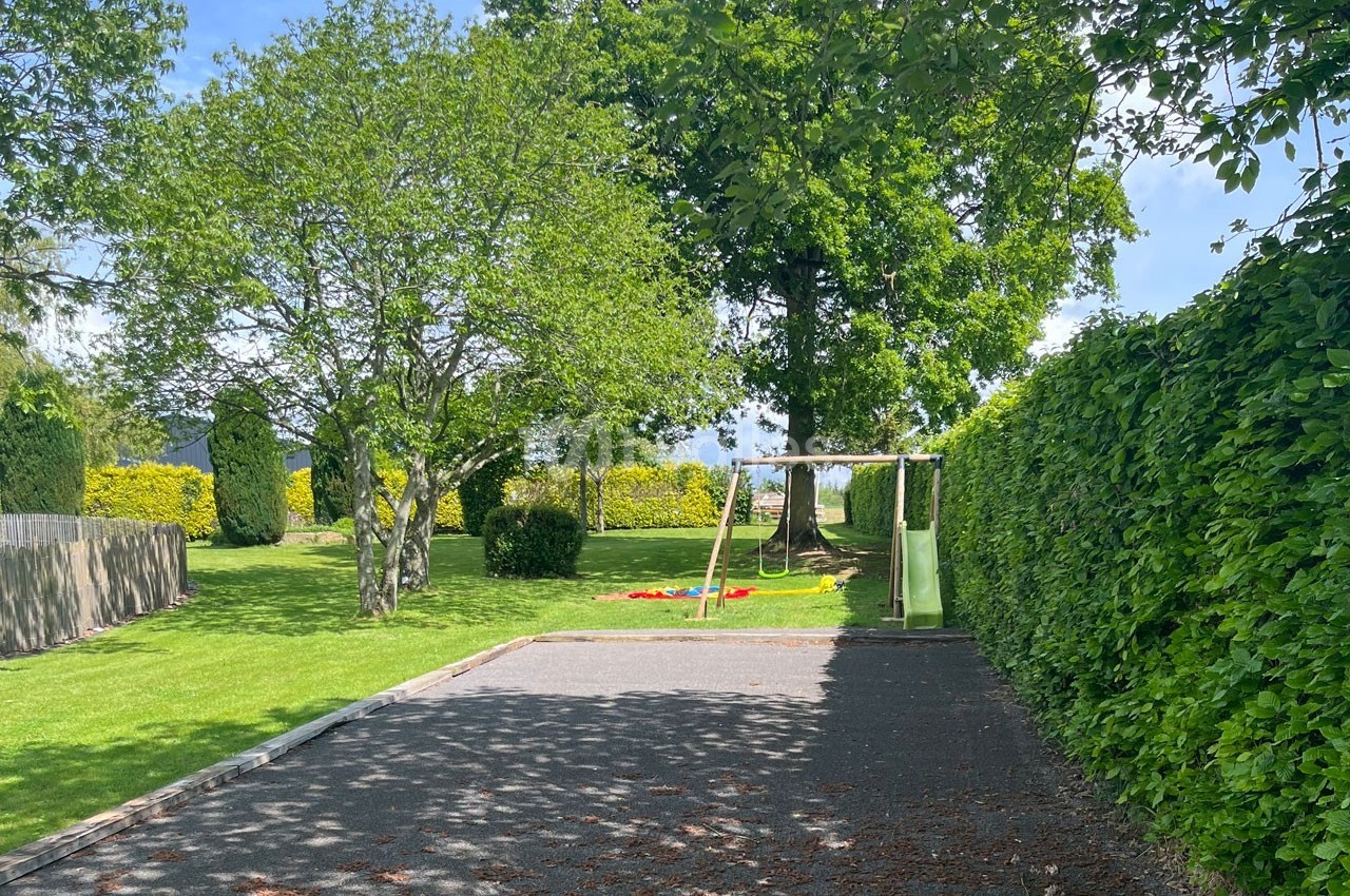 Allée ombragée menant à un jardin avec des arbres, une pelouse et une aire de jeux comprenant un toboggan.
