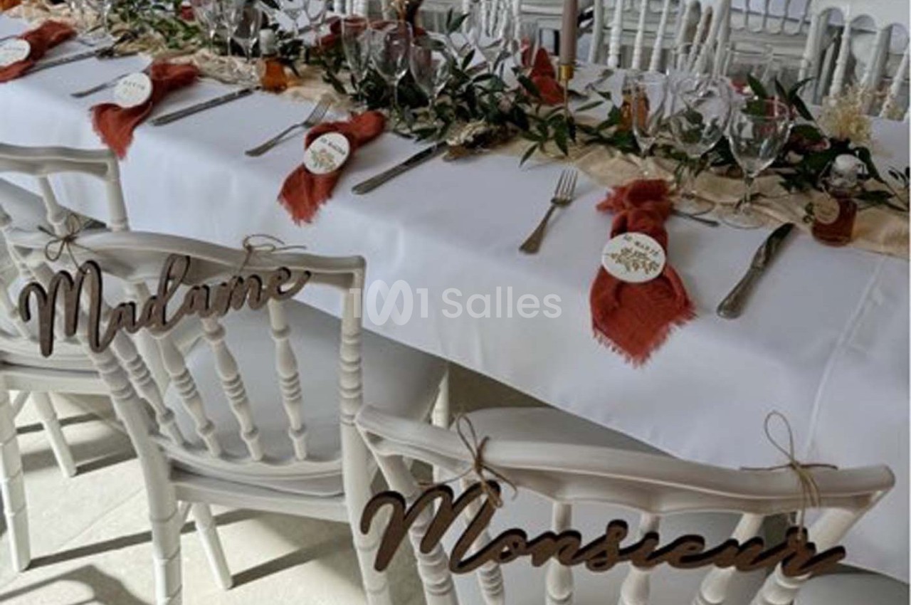 Table de réception décorée avec des couverts, serviettes rouges, feuillage et chaises marquées ’Madame’ et ’Monsieur’.