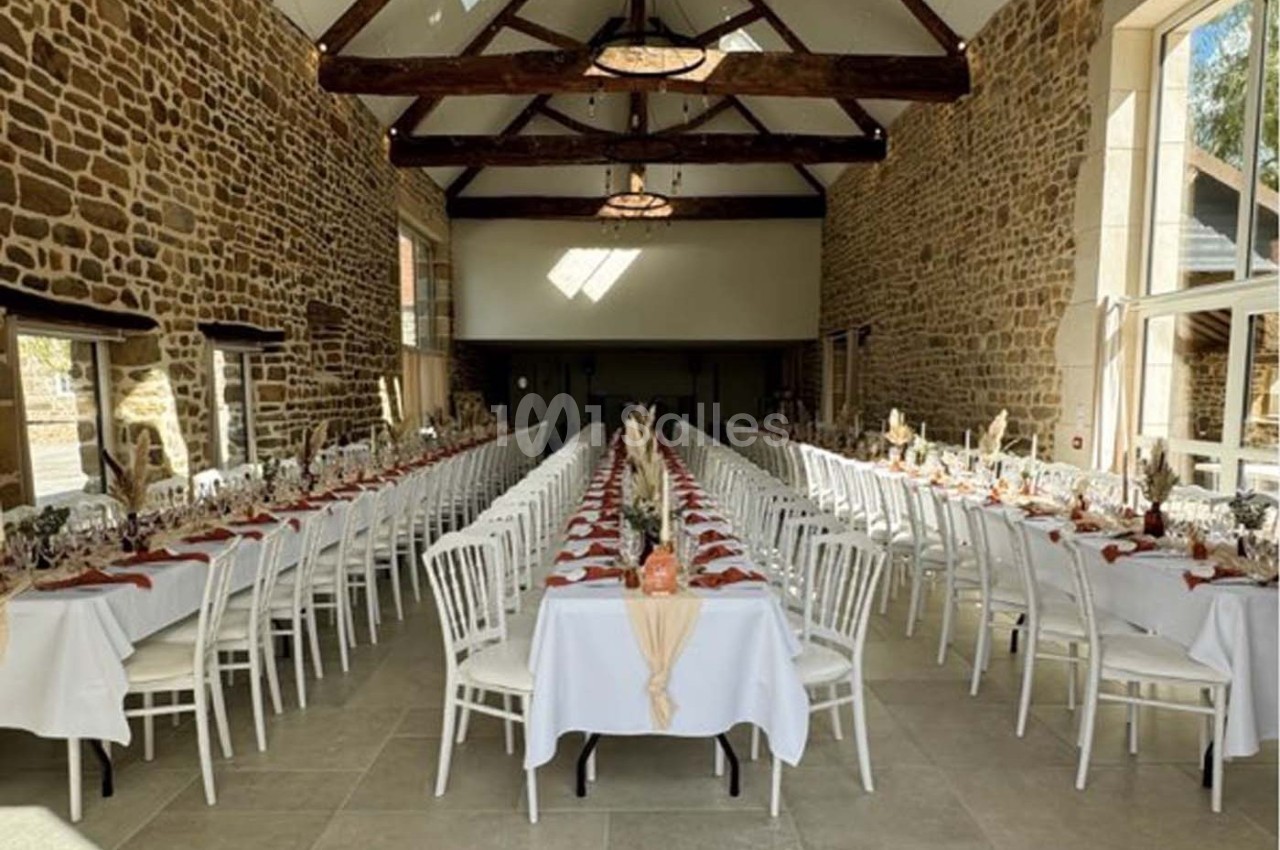 Salle de réception avec de longues tables dressées, nappes blanches, chaises alignées et murs en pierre apparente.