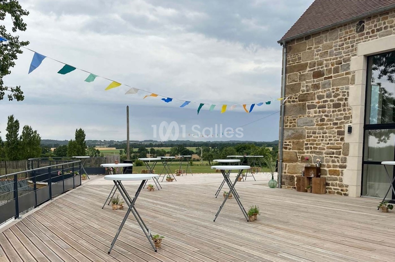 Terrasse en bois avec tables hautes, guirlandes colorées et vue sur un paysage champêtre sous un ciel nuageux.